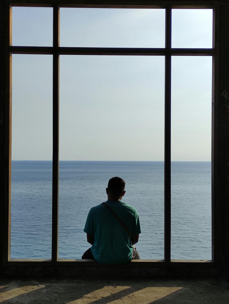 A lone man sits at a window overlooking the calm ocean, evoking solitude and reflection.
